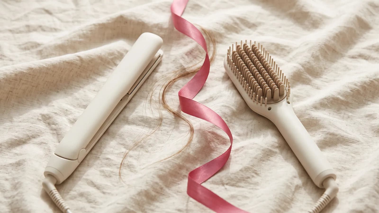 Un lisseur ivoire et une brosse lissante à picots posés côte à côte sur un lin crème, avec un ruban rose framboise et quelques mèches de cheveux