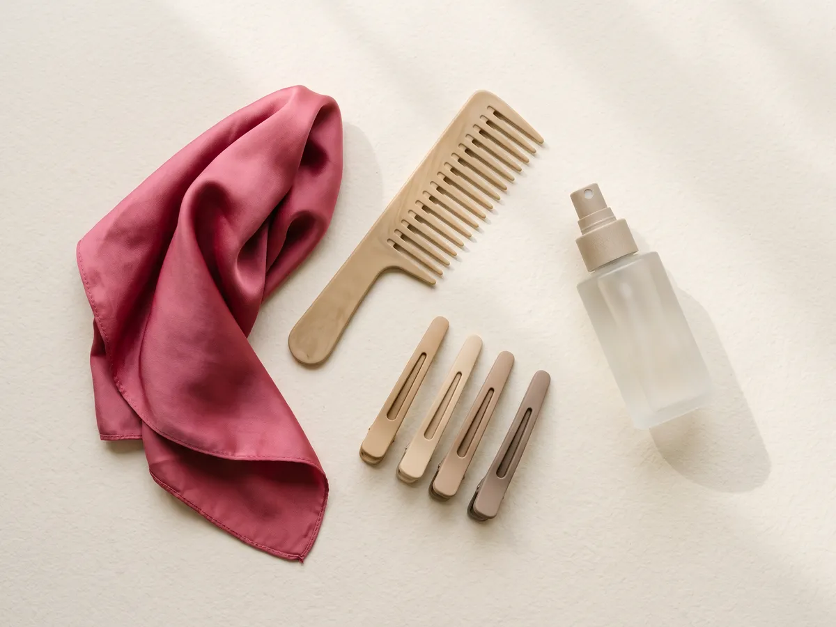 Peigne à dents larges, pinces de sectionnement, flacon sans étiquette et foulard en satin posés sur un fond crème